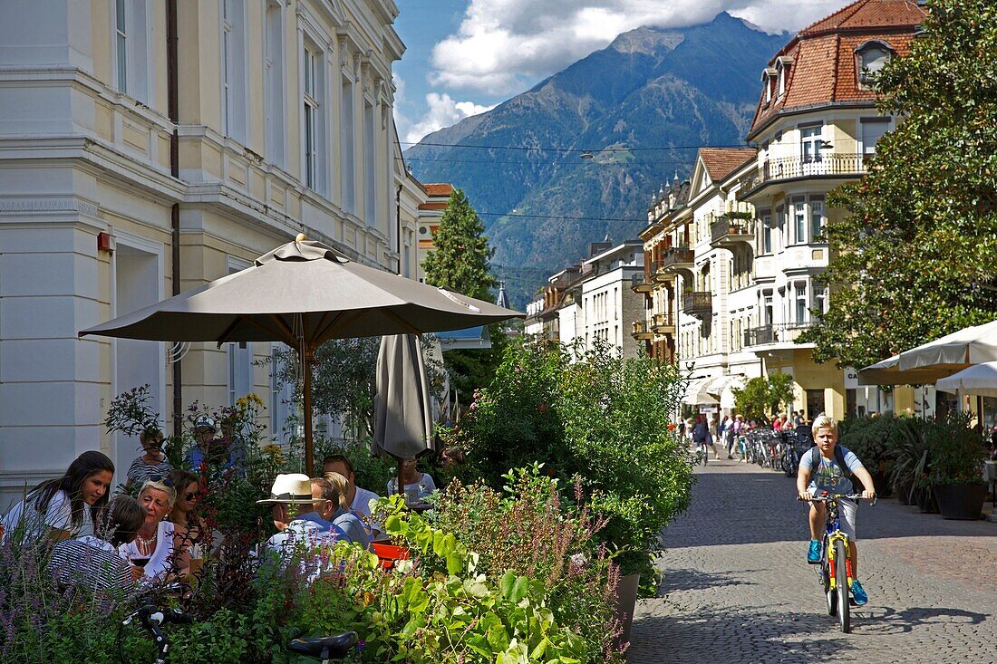 Italien, autonome Provinz Bozen, Meran, Kinder auf Fahrrädern und Menschen, die auf der Terrasse eines Cafés im Stadtzentrum sitzen, mit den grünen Bergen im Hintergrund