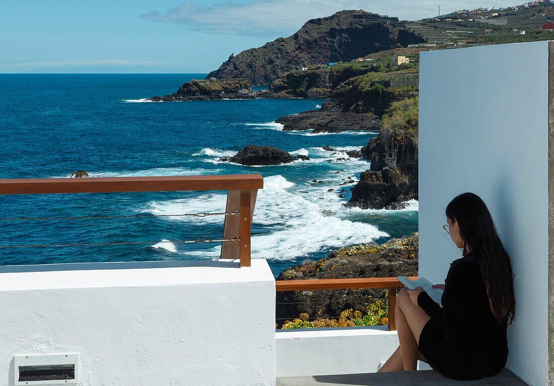 Spain, Canary Islands, La Palma, young and pretty woman sitting on a terrace by the sea, diving in the reading of a novel