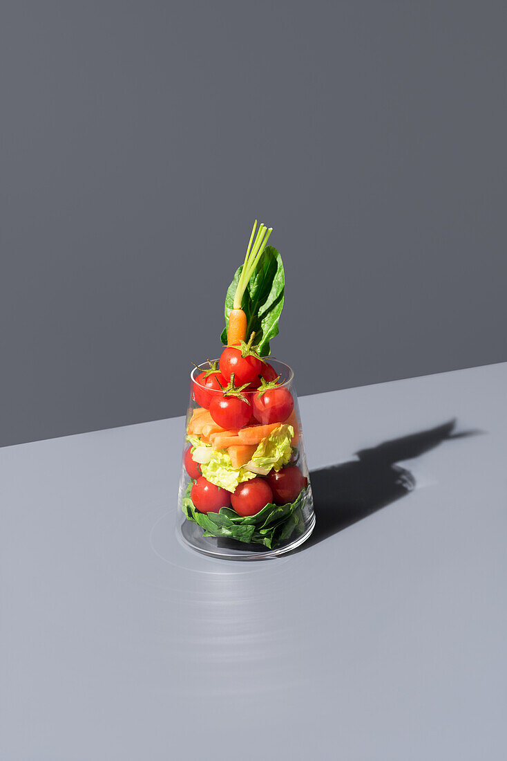 Geschichteter Salat, der in einem transparenten Glas vor grauem Hintergrund kunstvoll präsentiert wird, wobei frische Zutaten wie Tomaten, Karotten und Salat hervorgehoben werden.