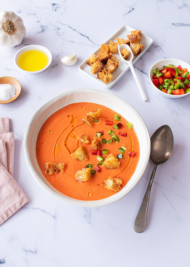 Blick von oben auf eine lebhafte Schüssel mit traditioneller spanischer Tomaten-Gazpacho-Suppe, serviert mit Knoblauch-Croutons und frischem, gehacktem Gemüse auf einer Marmor-Arbeitsplatte.