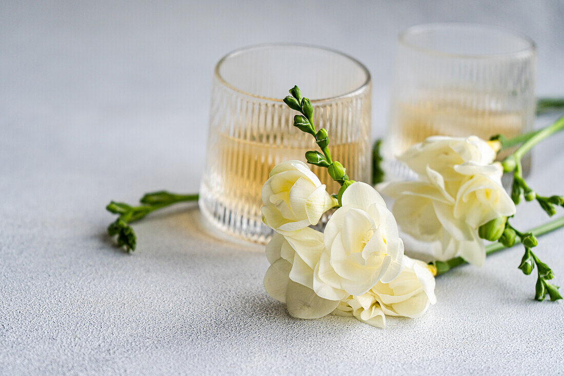 Ein raffiniertes Bild mit einem Pfirsich-Wodka-Cocktail in einem gerippten Glas, ergänzt durch zarte weiße Freesienblüten, vor einem strukturierten grauen Hintergrund.