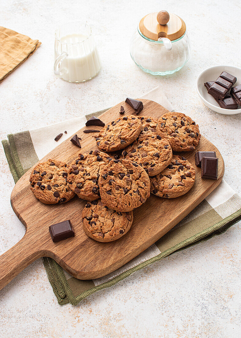 Freshly baked chocolate chip cookies laid out on a wooden serving board, accompanied by a jug of milk and pieces of dark chocolate