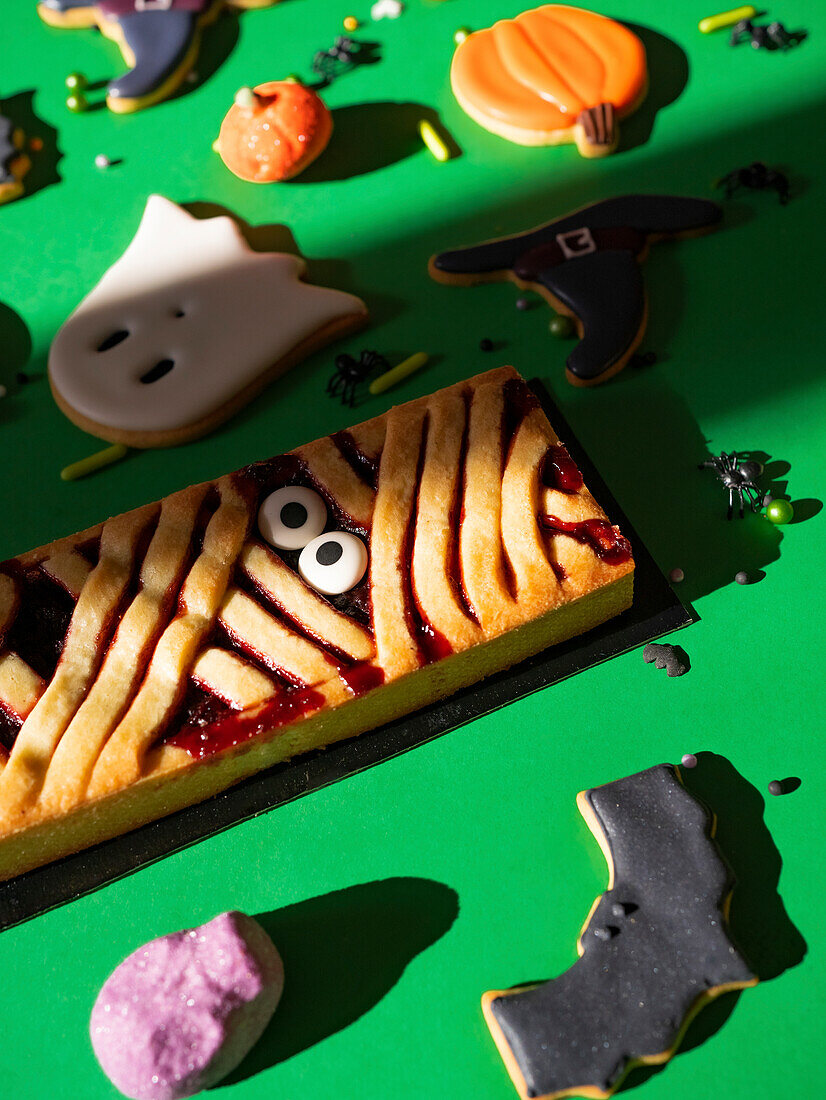 A selection of homemade Halloween cookies in spooky shapes and a large themed cookie with fruit filling and decorative eyes, on a green background with little festive touches