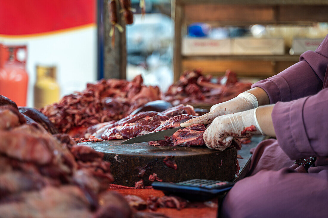 Unkenntlich gemachter Marktverkäufer in lila Kleidung schneidet fachmännisch frisches Rindfleisch auf einem großen Schneidebrett auf einem lebhaften Straßenmarkt in Bangkok, der die lebhafte lokale Lebensmittelszene zeigt.