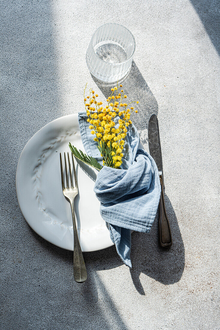 Draufsicht auf einen frisch gedeckten Tisch im Freien mit einem weißen, geprägten Teller, Silberbesteck, einer blauen Leinenserviette und einem Zweig mit leuchtend gelben Mimosenblüten