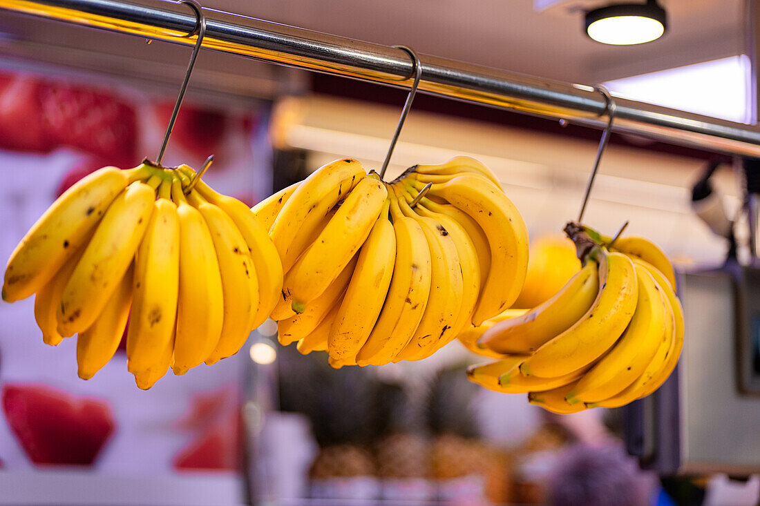 Ripe yellow bananas hang from a metal bar in a bustling market setting with a hint of fruit stalls in the background