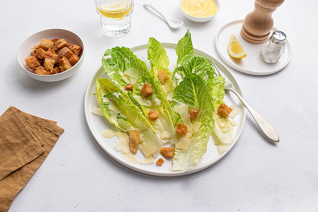 Draufsicht auf einen klassischen Caesar-Salat mit knackigem Römersalat, knusprigen Croutons, gehobeltem Parmesankäse und cremigem Dressing, serviert auf einem weißen Teller auf weißem Hintergrund.