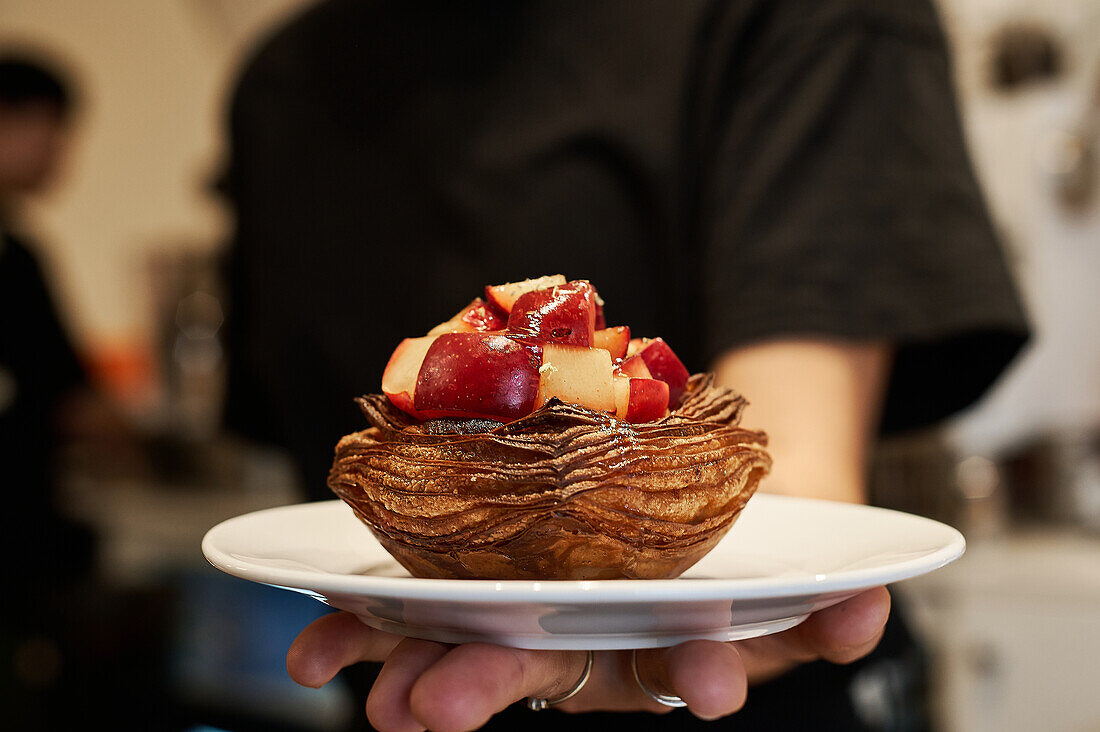 Unrecognizable waiter serving a delectable pastry topped with fresh fruit, presented on a white plate with a blurred background.