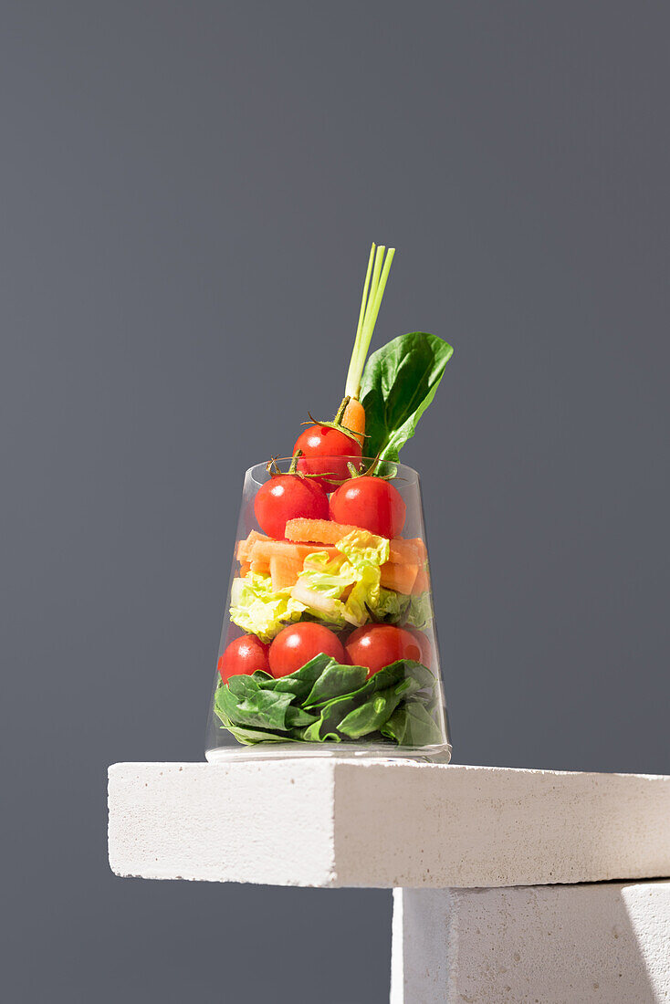 Frischer und farbenfroher geschichteter Salat, serviert in einem transparenten Glas, elegant präsentiert auf grauem Hintergrund. Gesunde Mahlzeit mit einer Reihe von Gemüsesorten wie Tomaten, Spinat und Karotten