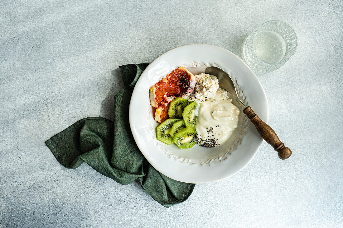 Blick von oben auf ein ausgewogenes Frühstücksset mit einer weißen Schale mit Hüttenkäse, Naturjoghurt und Chiasamen, dazu Kiwi und sizilianische Orangenscheiben