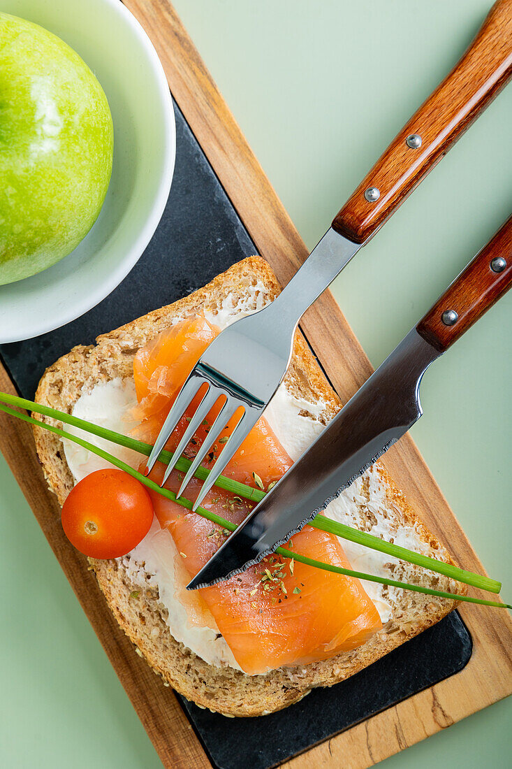 Top view of a nutritious breakfast featuring salmon on whole grain bread, garnished with fresh chives and cherry tomato, alongside a green apple.