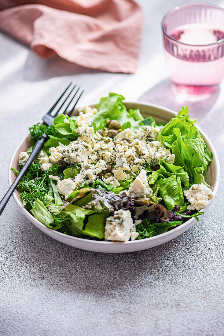 Frischer und gesunder Kräutersalat mit Kopfsalat, grünen Zwiebeln, Petersilie, Estragon, blauem Brie-Käse und fermentierten Kapern, beträufelt mit Olivenöl, serviert in einer Keramikschale neben einem Glas Wasser