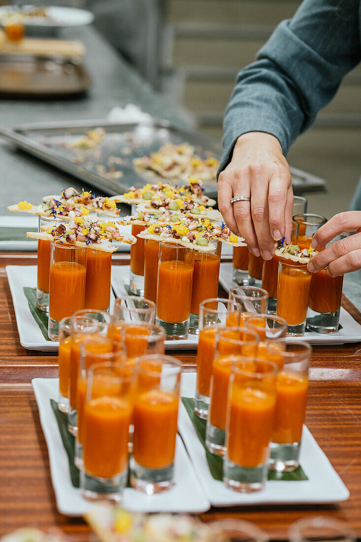 Anonymous chef hand adorns glasses of vibrant orange soup with seasoned crackers as a finishing touch before service