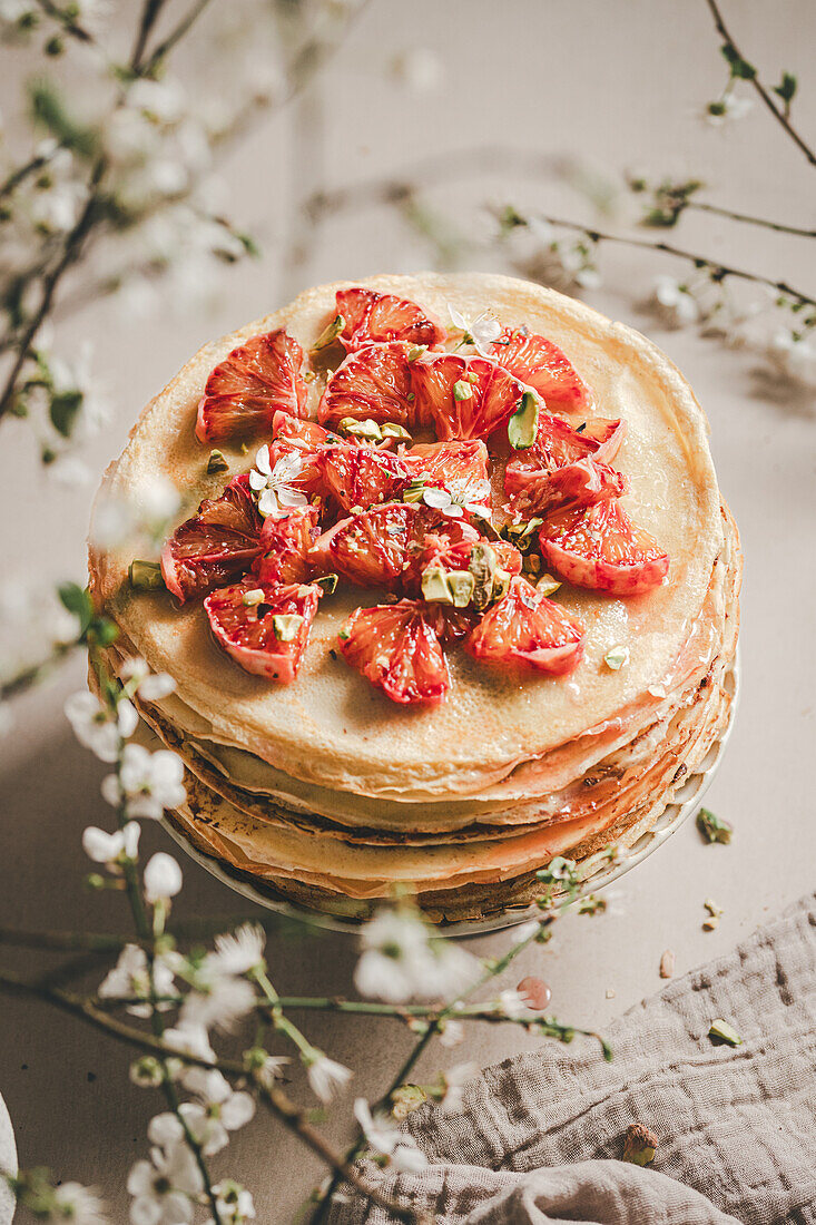 Ein Stapel hausgemachter Crêpes, verziert mit frischen Erdbeerscheiben und Pistazien, vor einem zarten Blumenhintergrund