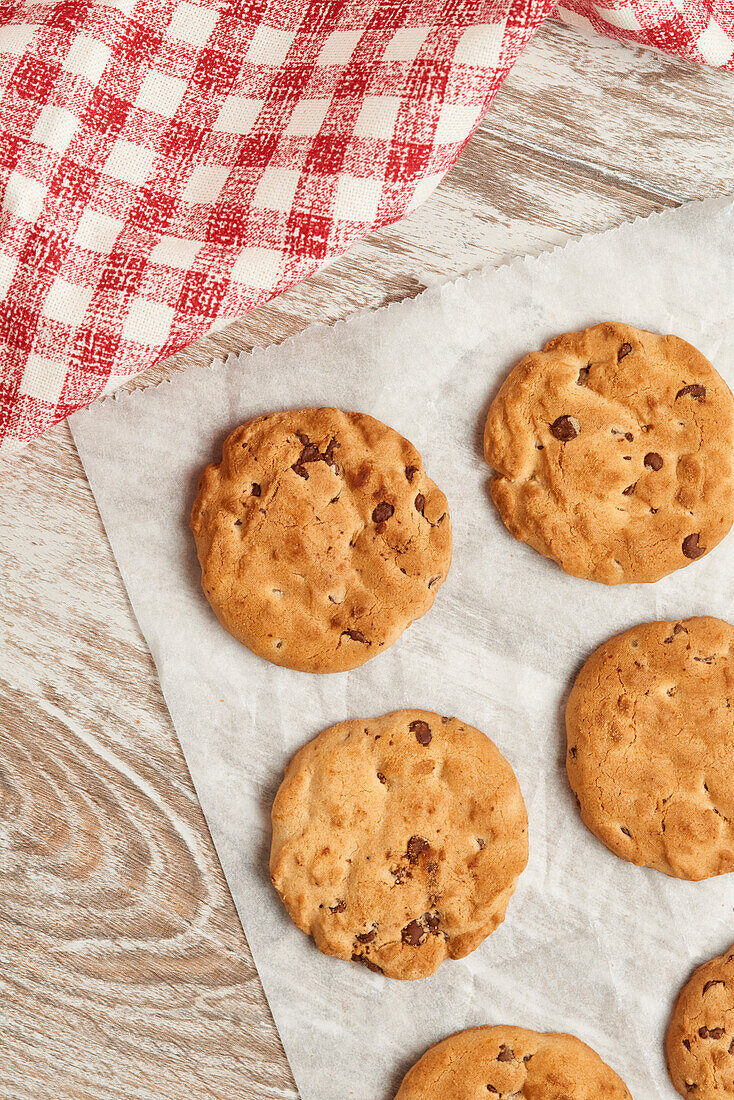 Chocolate chip cookies laid out on parchment paper with a classic gingham cloth beside them, captured from a top-down perspective