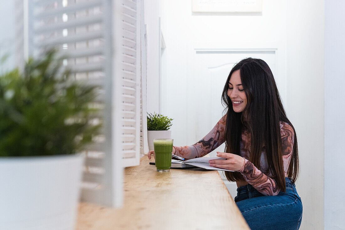 Smiling female sitting at counter with cocktail in bar and writing in notepad while looking at camera