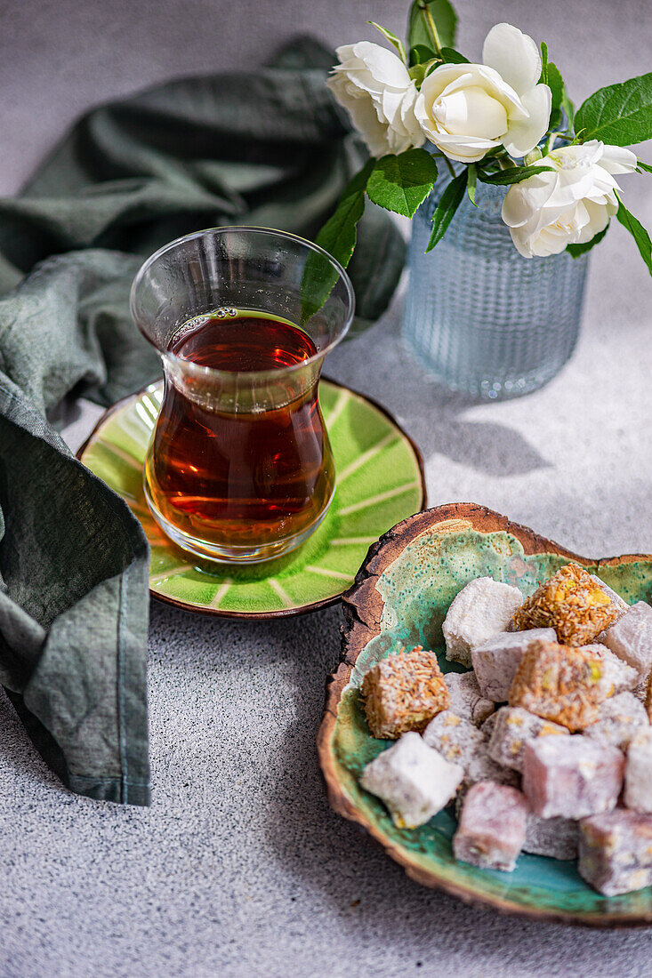 Assorted traditional Turkish delights on a ceramic plate beside a glass of black tea, with a vase of white roses in the background