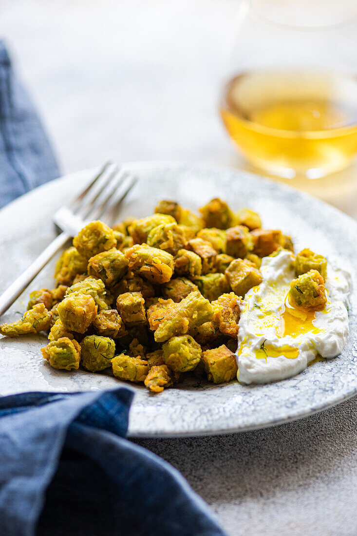 Ein Gericht aus gebratener Bamia, großzügig mit einer Sauerrahmsauce mit Dill und Knoblauch übergossen, wird neben einem Glas Wein präsentiert und verkörpert die Essenz der mediterranen Küche