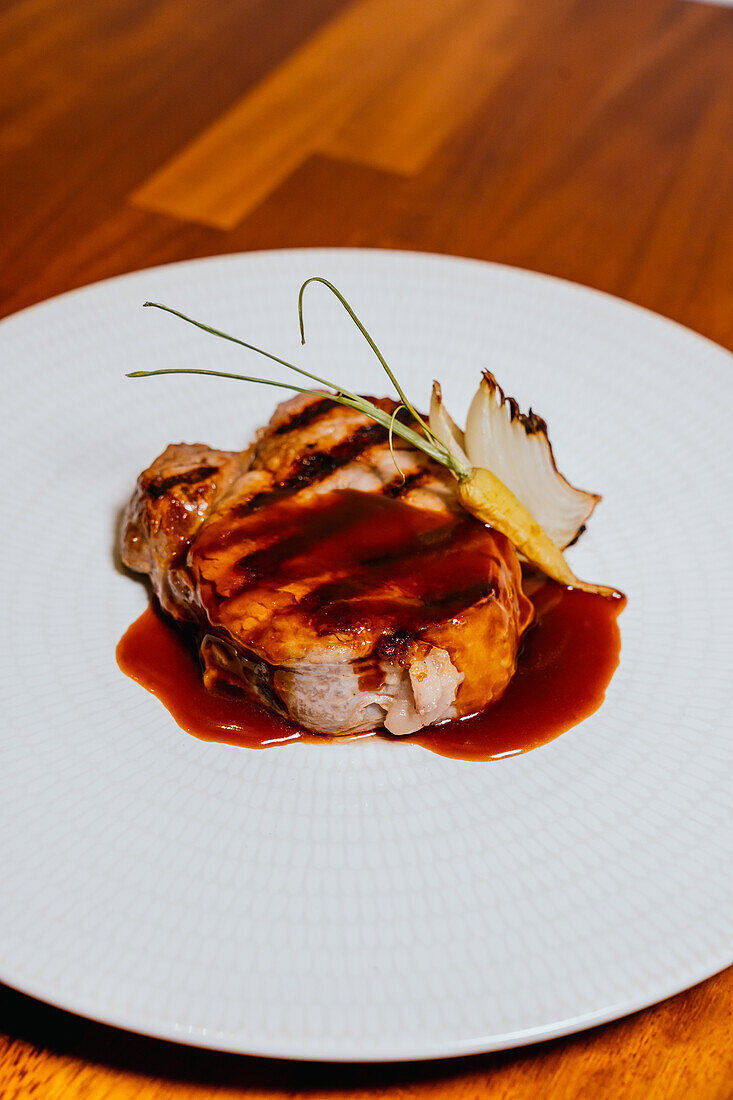 A succulent pork steak drizzled with a rich glaze, garnished with an onion and delicate greens, served on a textured white plate against a wooden background