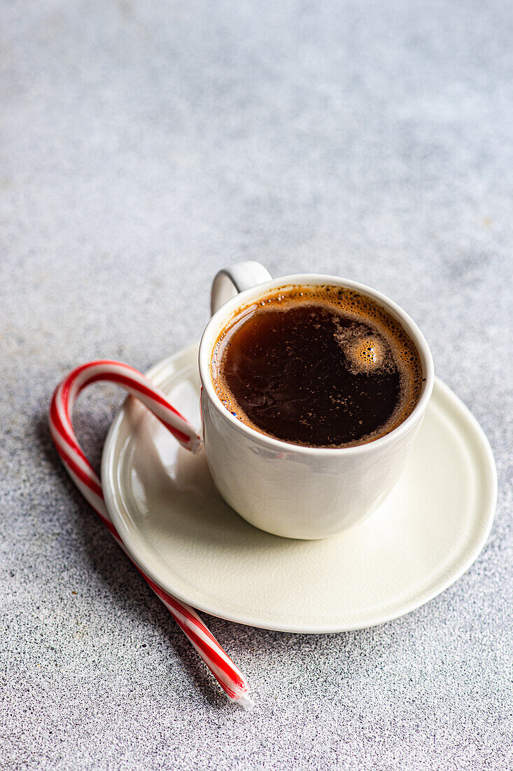 A cozy morning scene featuring a white mug of hot coffee paired with a red and white candy cane on a speckled grey surface.