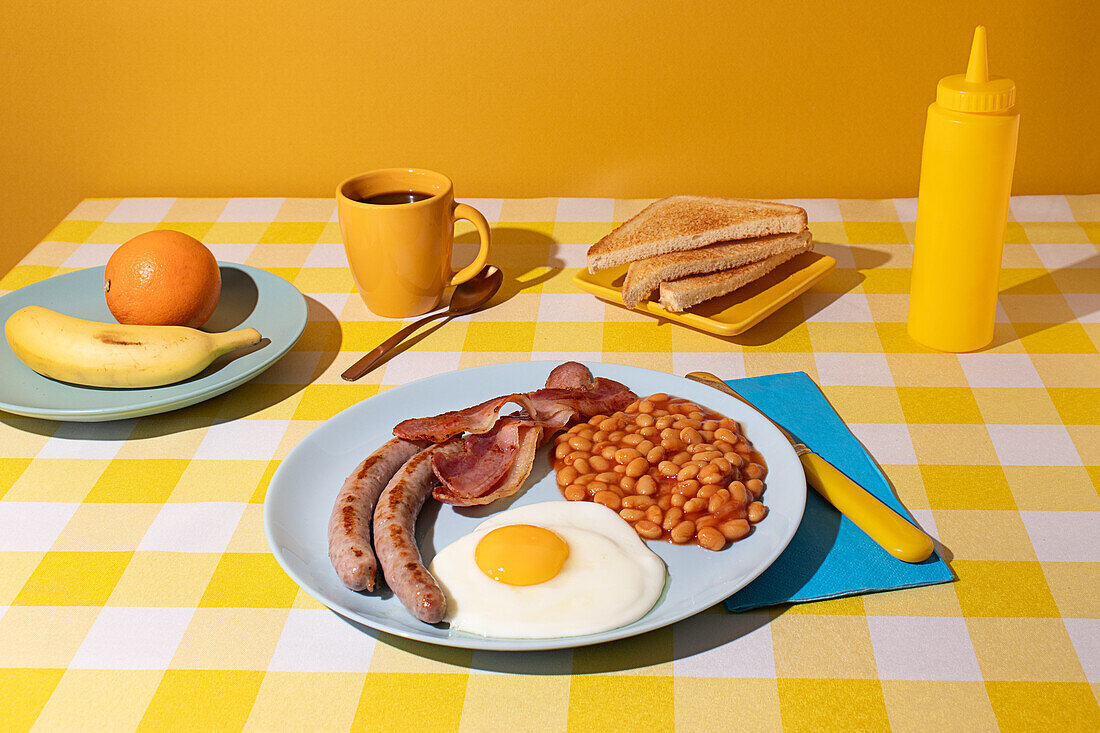 Yellow coloured Tablecloth and background with a delicious full English Breakfast, egg, beans, sausages and bacon, some toasted bread slices, fruit and a cup of coffee