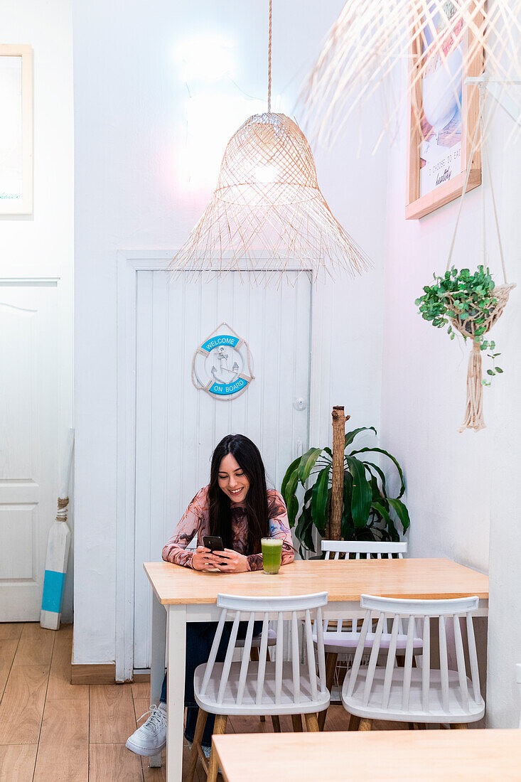 Positive female sitting at table with smoothie and browsing mobile phone while chilling at weekend in bar