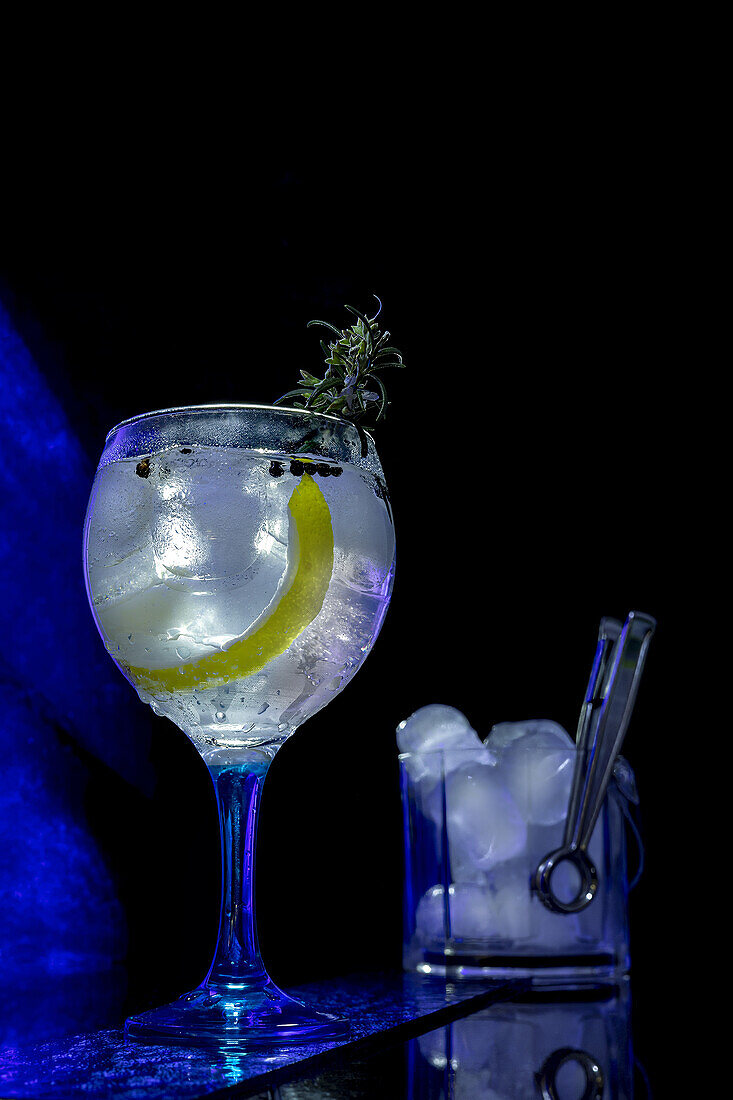 Classic gin tonic cocktail in a balloon glass with ice and a slice of lime, tongs, and ice cubes visible on the side