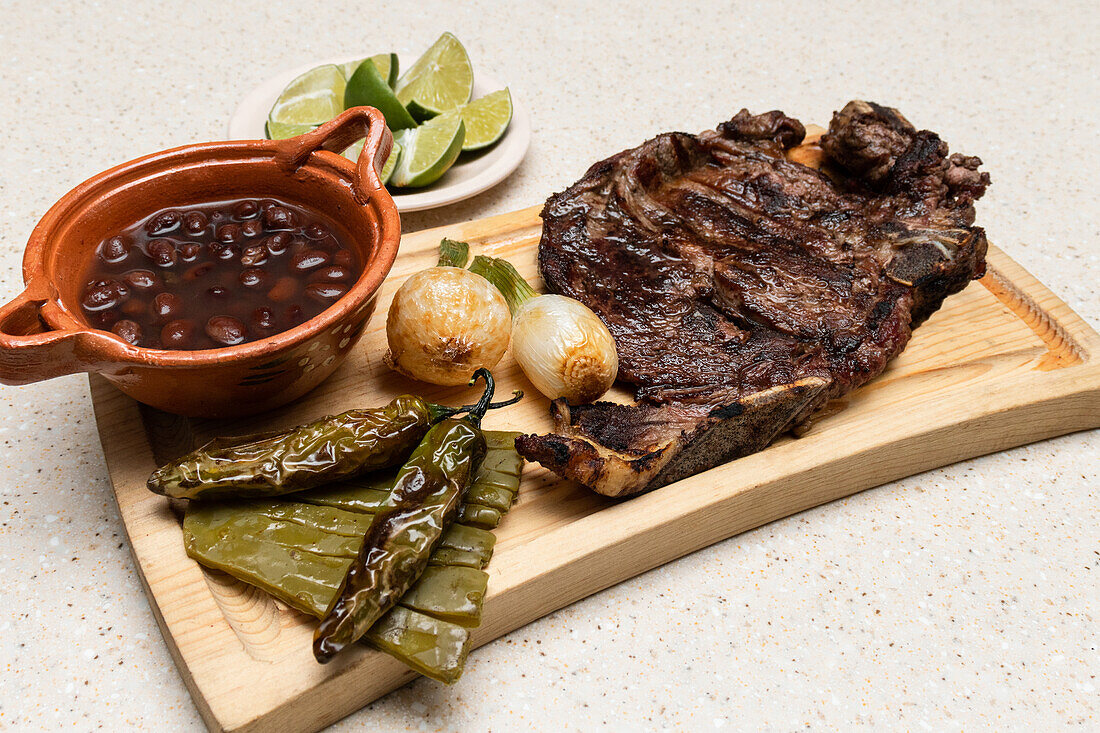 Appetizing display of traditional Mexican cuisine, featuring char-grilled ribs on a wooden board, accompanied by roasted chilies, onions, a bowl of black beans, and slices of lime.