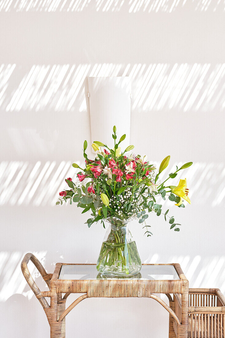 Ein wunderschönes Gesteck aus frischen Blumen in einer durchsichtigen Vase steht auf einem Rattantisch, während das Sonnenlicht gestreifte Schatten auf eine weiße Wand wirft