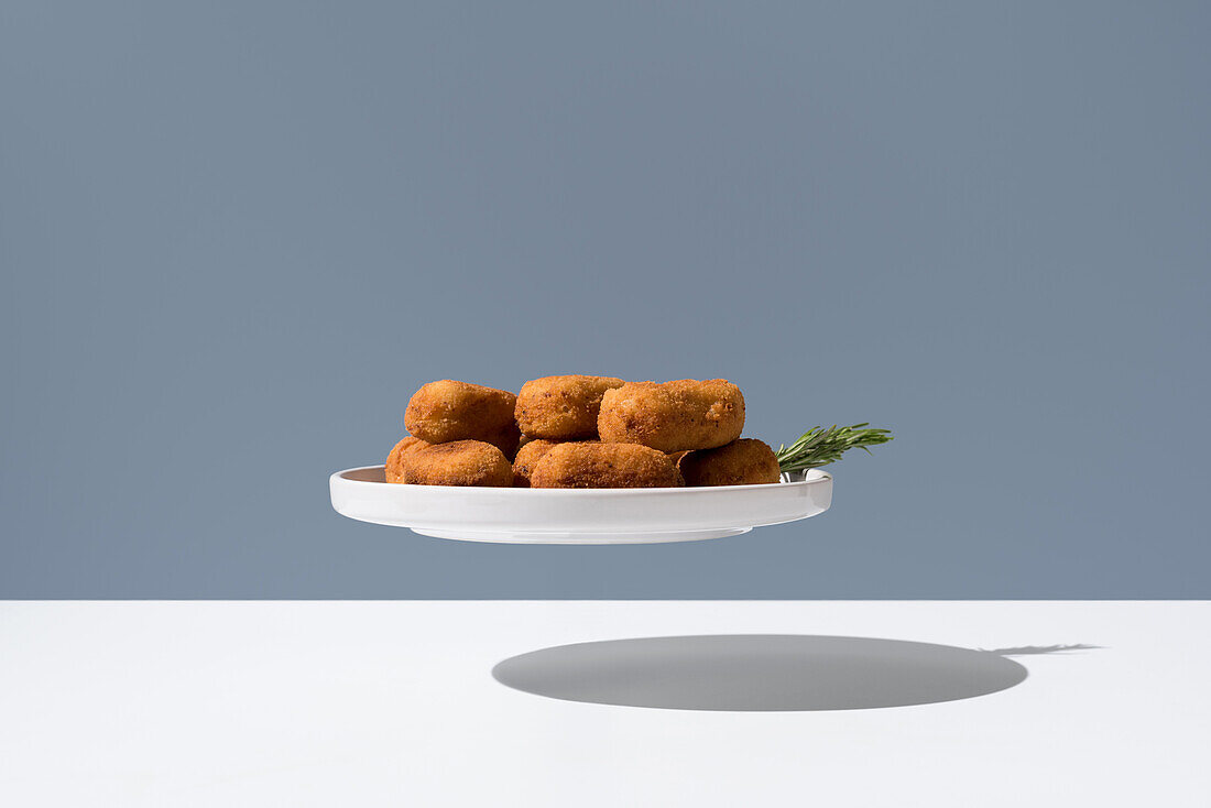 Levitating plate of crispy croquettes garnished with fresh herb on a gray background with shadow below