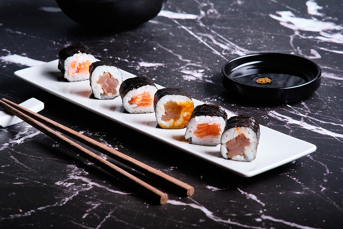 Assorted sushi rolls served on a white plate with soy sauce and chopsticks on a dark marble background.