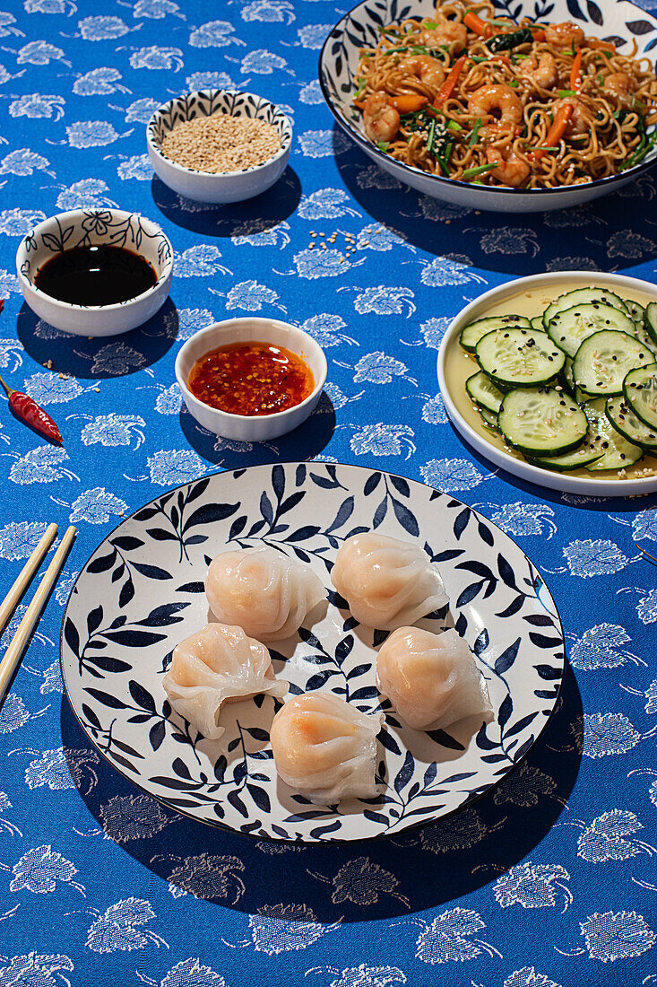 Draufsicht auf ein traditionelles chinesisches Essensarrangement mit Krabben-Nudeln, Knödeln, Gurkenscheiben und Soßen auf einem blau gemusterten Tischtuch, beleuchtet von natürlichem Sonnenlicht