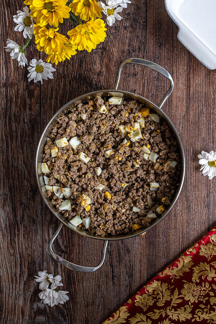 Top view of a freshly prepared homemade meat pie filling in a stainless steel pan, featuring ground meat, potatoes, and boiled eggs on a rustic wooden surface, surrounded by yellow and white flowers.