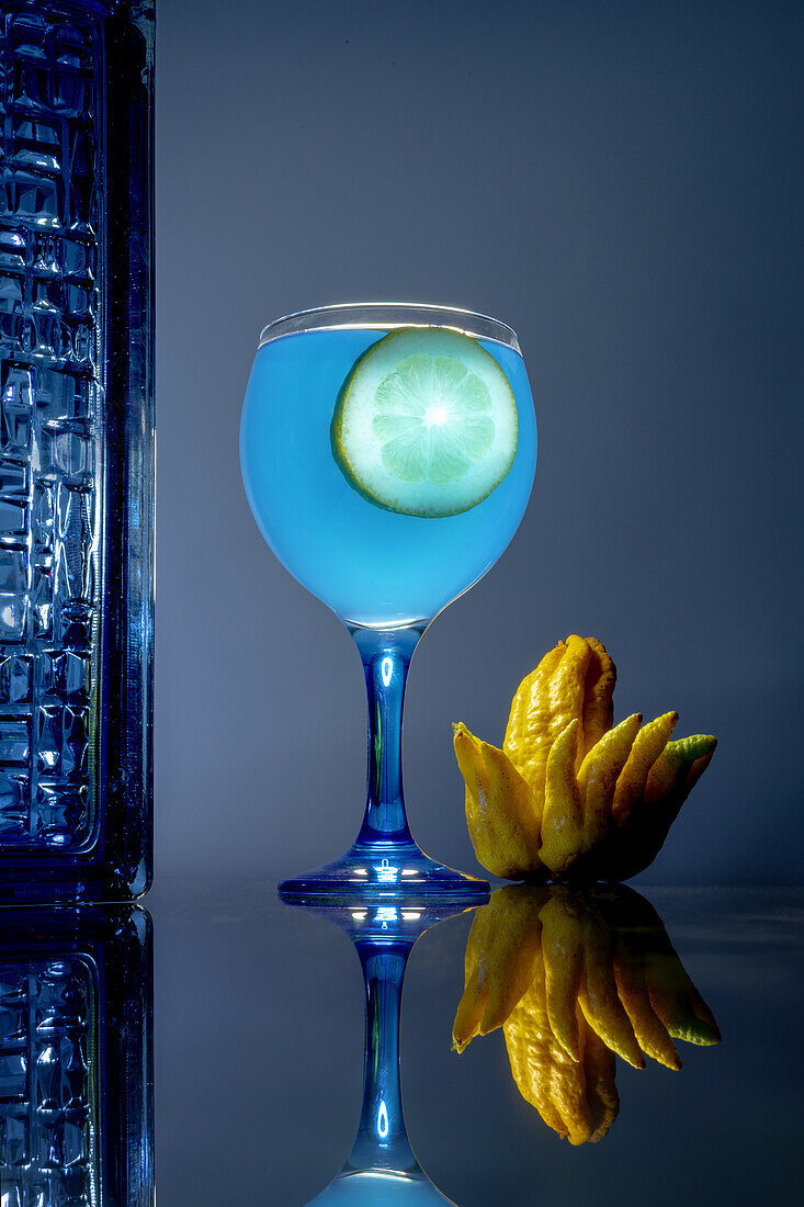 Ein leuchtend blauer Cocktail mit einer Limettenscheibe und einer Zitrusfrucht in der Hand eines Buddhas auf einer spiegelnden Oberfläche