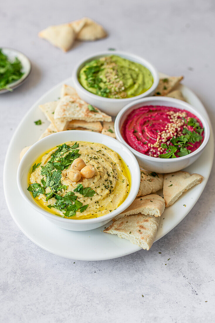 Drei verschiedene Hummus-Variationen, klassisch, mit Rüben und Avocado, jeweils mit frischen Kräutern garniert und mit Fladenbrot serviert, werden in einer bunten Auslage präsentiert.