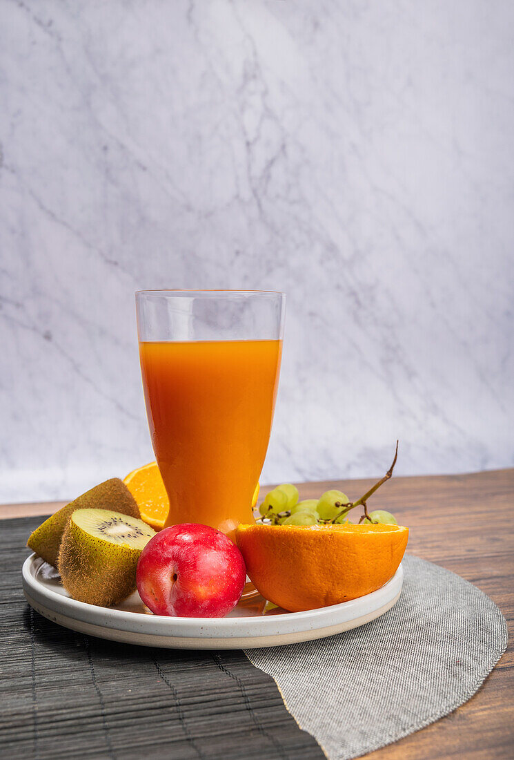 Eine bunte Auswahl an frischem Obst, darunter Kiwi, Orangen, Äpfel und Trauben, sowie ein Glas Orangensaft. Alles ist auf einem runden Teller vor einem Marmorhintergrund mit einer rustikalen Holzoberfläche angerichtet.