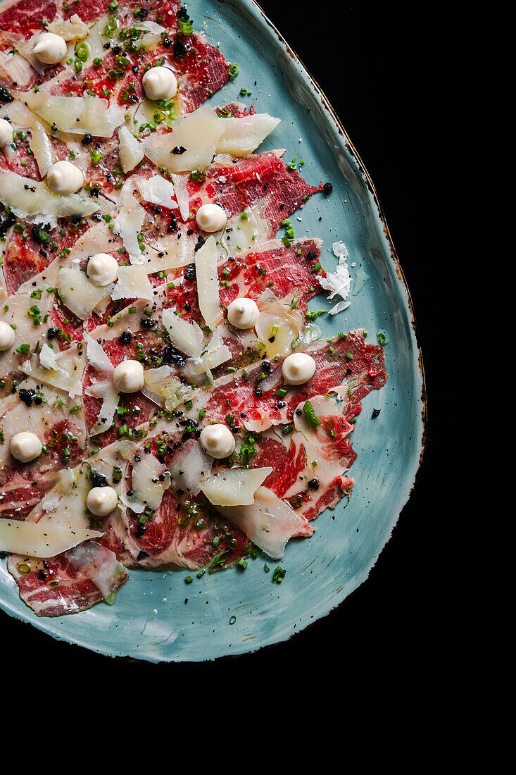 From above exquisite beef carpaccio adorned with shaved parmesan, garnished with herb sprinkles and decorative edible pearls on a ceramic plate.