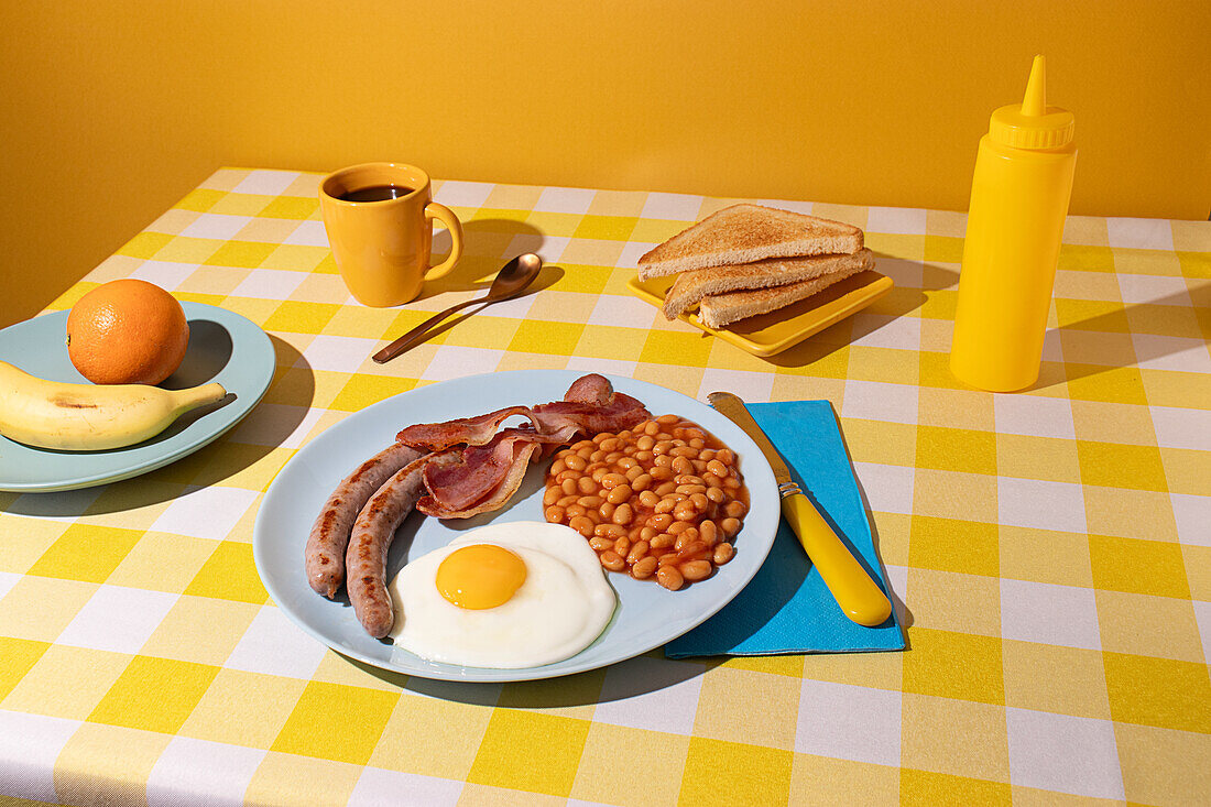 Yellow coloured Tablecloth with a delicious full English Breakfast, egg, beans, sausages and bacon, some toasted bread slices, fruit and a cup of coffee