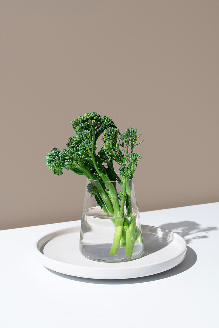 Fresh broccolini stalks, also known as baby broccoli or tenderstem, displayed in a clear glass jar filled with water, against a minimalist beige background.