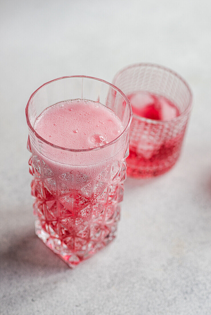 Zwei strukturierte Gläser mit einem erfrischenden Kirsch-Wodka-Tonic-Cocktail mit Sprudel und Eiswürfeln vor einem weichen Hintergrund.