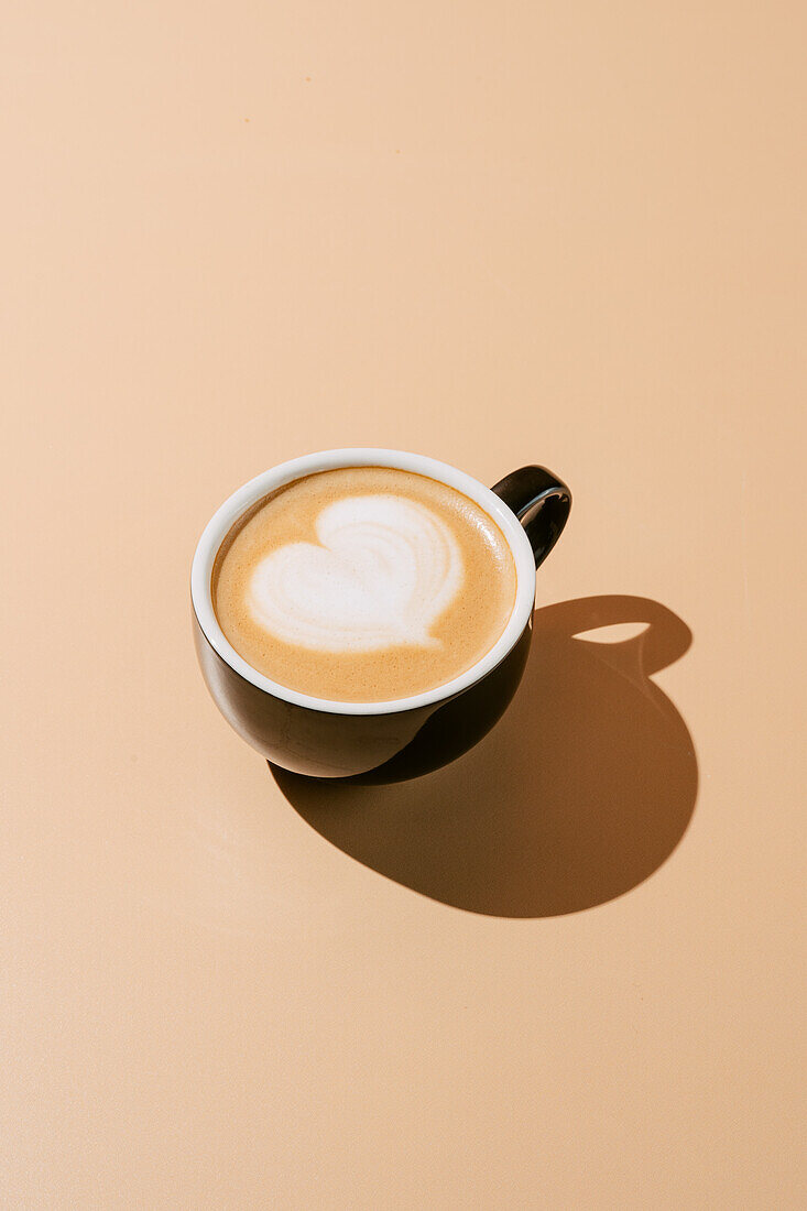 From above beautifully crafted cappuccino with heart-shaped latte art, presented in a classic ceramic cup cast with a soft shadow on a cream background.