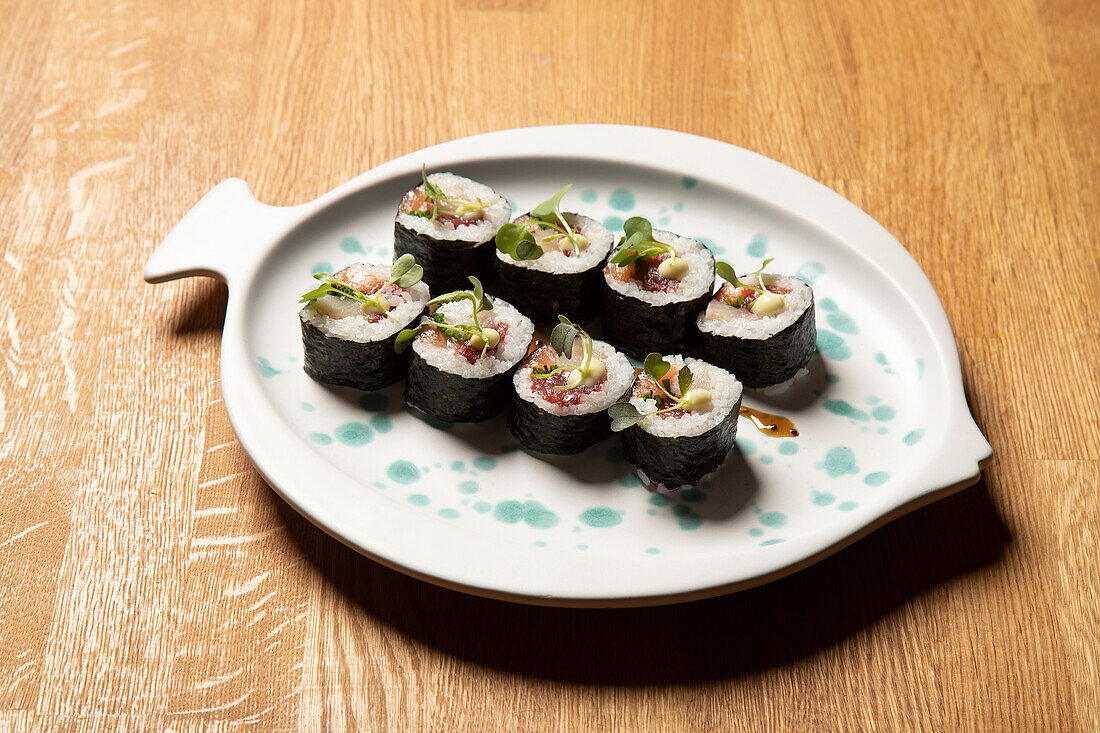 An artistic presentation of sushi rolls on a fish-shaped, white ceramic plate, garnished with green sprouts and drizzled with soy sauce, displayed on a wood-textured surface