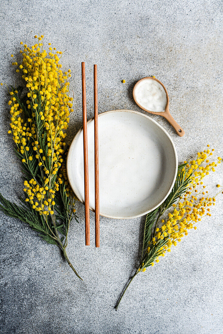 Draufsicht auf einen eleganten, minimalistisch gedeckten Tisch mit einem Keramikteller und Essstäbchen, akzentuiert durch das leuchtende Gelb von Mimosenblüten vor einem strukturierten grauen Hintergrund