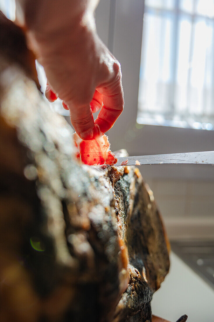 A close-up image capturing a hand carefully slicing thin pieces off a Serrano Ham leg, basked in natural light near a window