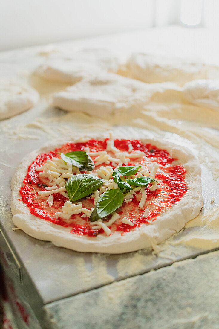 An anonymous chef prepares a pizza base topped with tomato sauce, cheese, and fresh basil, indicative of pizzeria craftsmanship