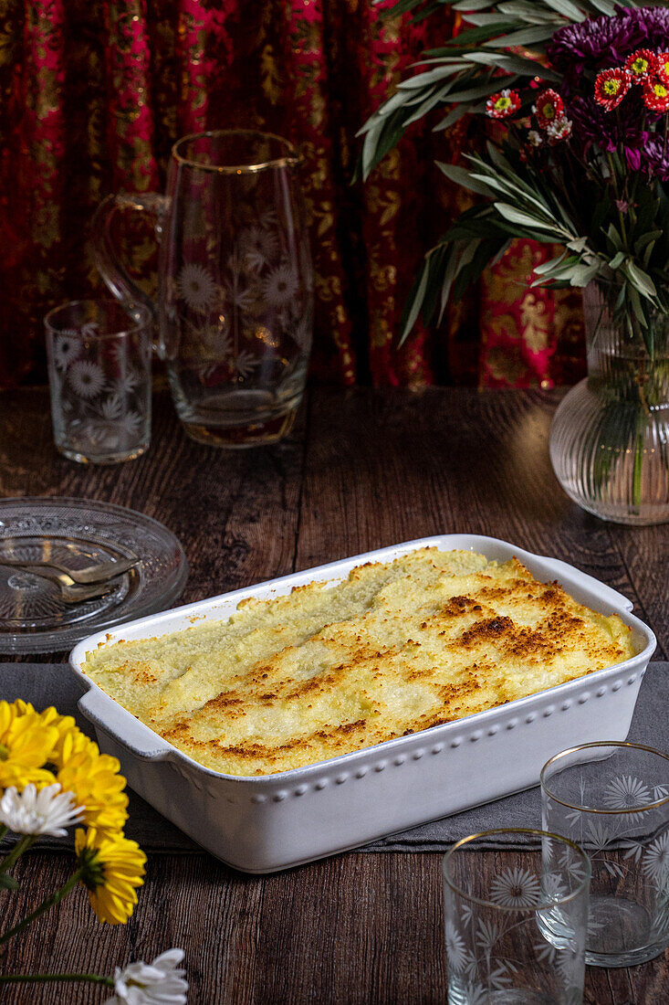 Köstliche hausgemachte Fleischpastete in einer weißen Auflaufform, serviert auf einem Holztisch. Das Gericht ist von eleganten Gläsern und bunten Blumen umgeben, die eine gemütliche, einladende Speiseszene schaffen.