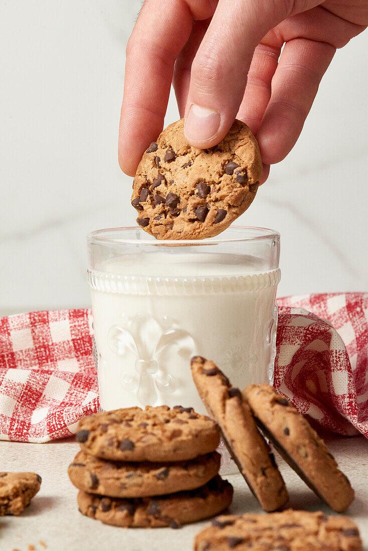 A hand is captured placing a chocolate chip cookie atop a full glass of milk, with a stack of cookies and a checkered napkin beside it