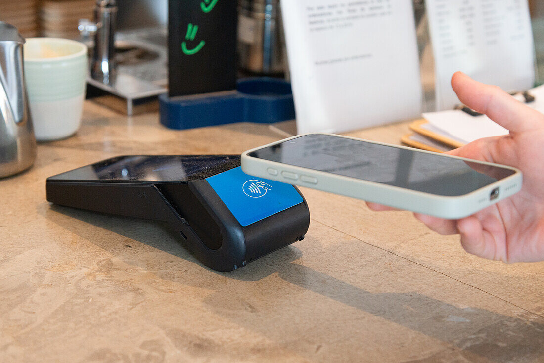 Cropped unrecognizable image of a hand holding a smartphone above a payment device to make a contactless purchase at a coffee shop.