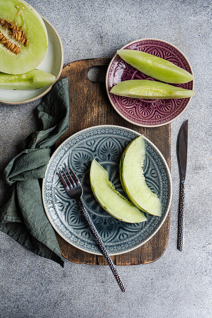 Draufsicht auf frisch geschnittene Bio-Mini-Melonen, die elegant auf dekorativen Tellern angerichtet sind, begleitet von einer dunklen Metallgabel und einem rustikalen Holzbrett, bereit für ein erfrischendes Mittagessen.