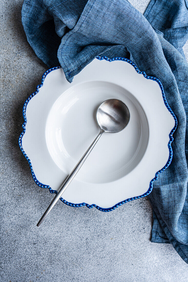 An elegant white plate with blue edges and a silver spoon, accompanied by a denim blue napkin on a textured surface